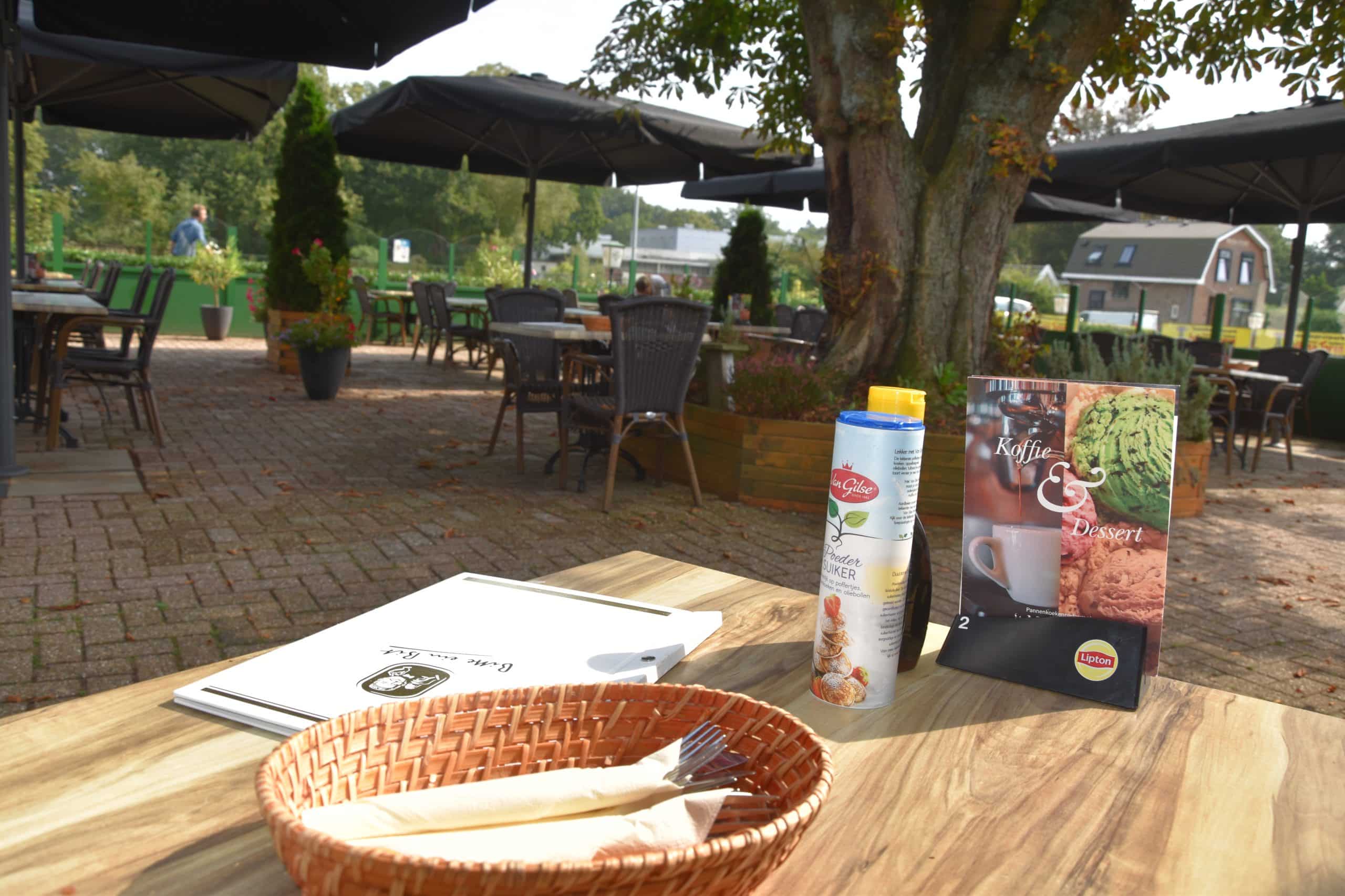 Een houten tafel op het terras met broodmandje, poedersuiker en de kaart voor koffie en dessert.