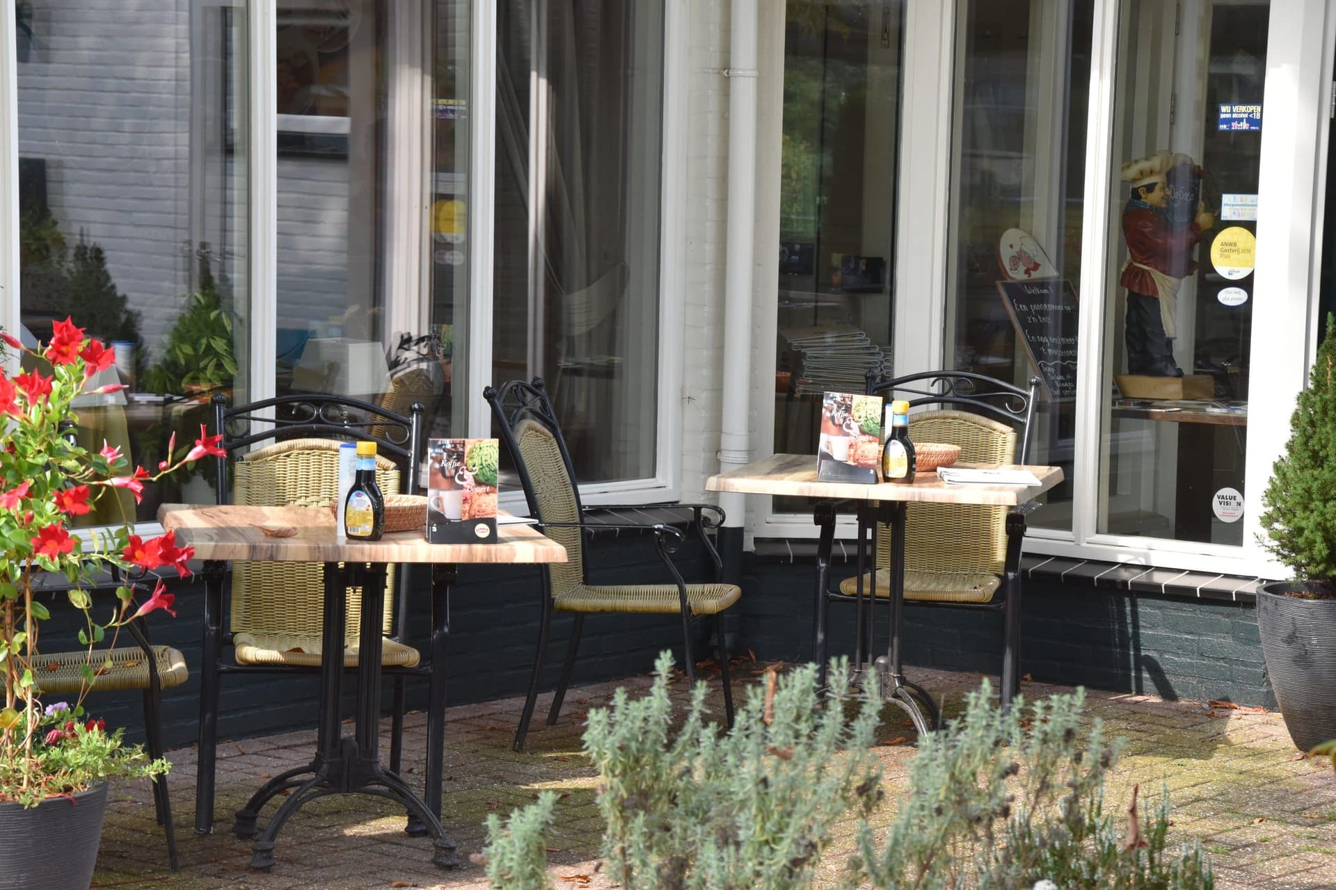 Twee tafeltjes met rieten stoelen in de zon voor de ramen van het restaurant.