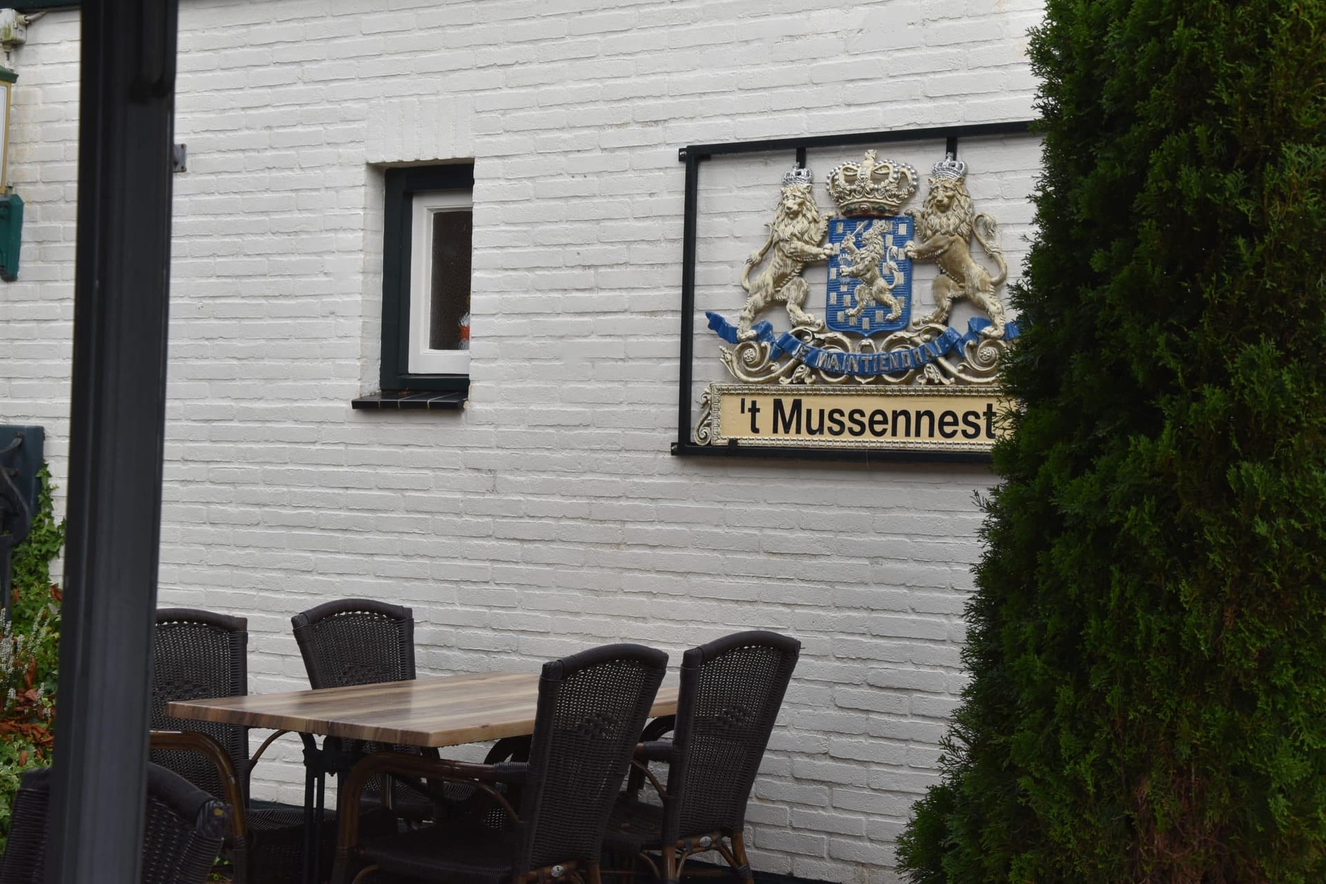 Het naambord van 't Mussennest met wapen op de wit geschilderde bakstenen gevel, naast een gedekte tafel.