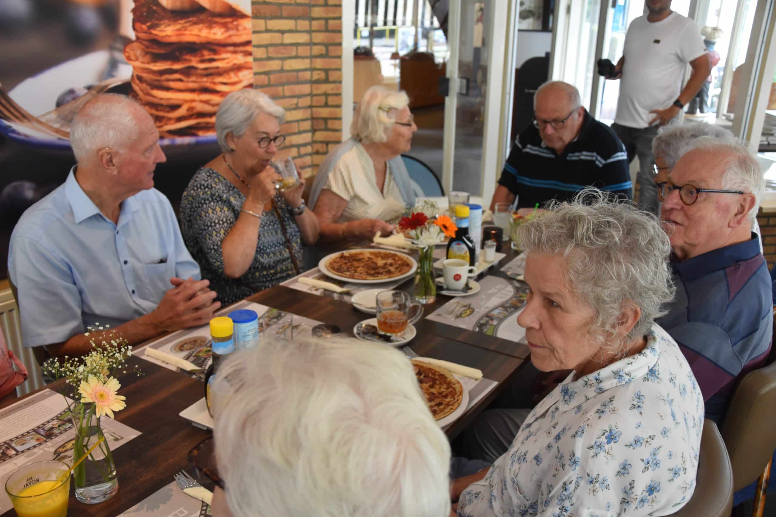 Gasten aan een lange tafel met pannenkoeken, koffie en bloemen in het restaurant.