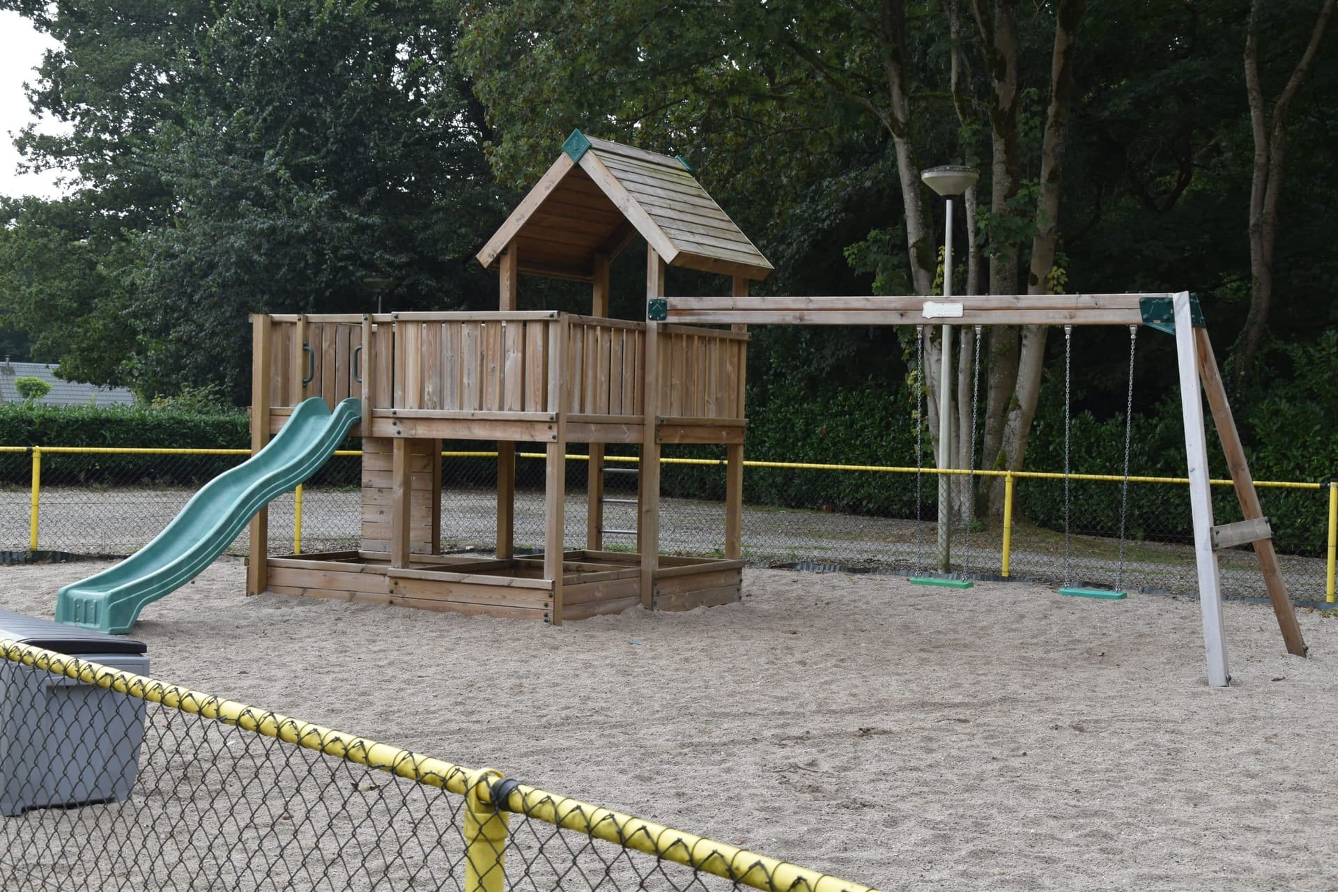 De omheinde buitenspeeltuin met houten speelhuis, glijbaan en schommels in het zand.